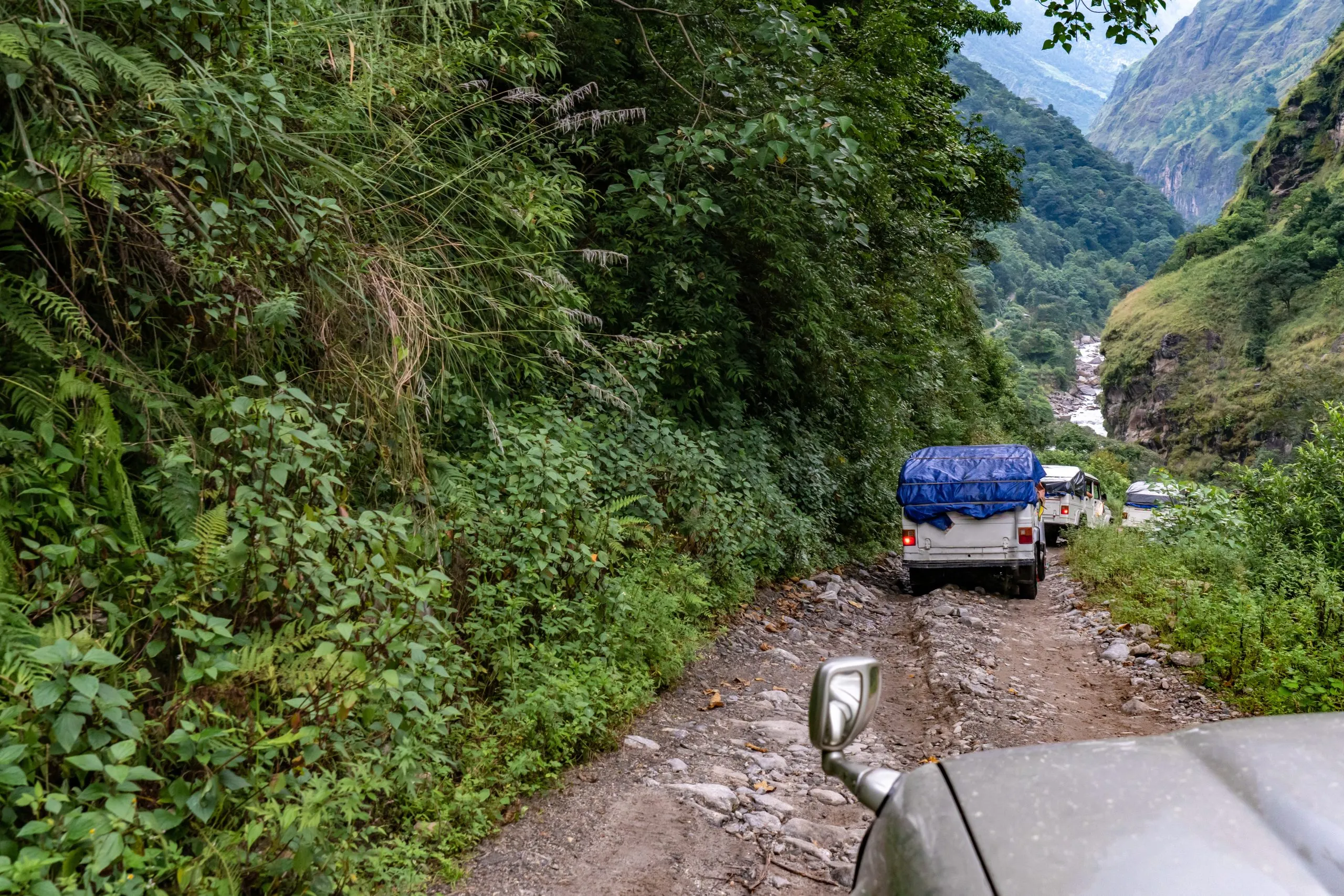 A column of 4wd vehicles carrying loads descending a dirt and rock path into a deep canyon lined with a river, Manang Road, Nepal